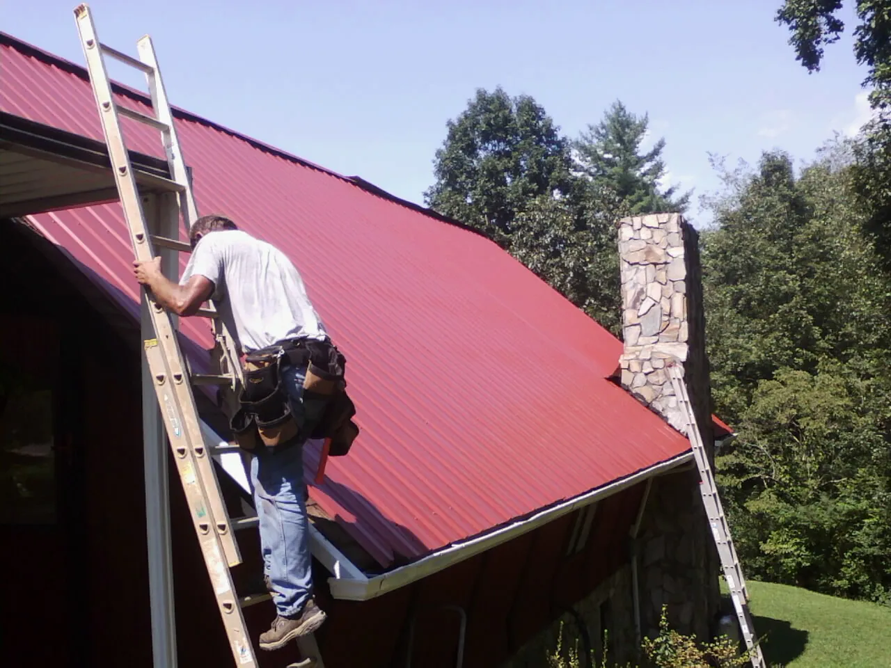 Roofer on a ladder beside a red metal roof and stone chimney — Soffit & Fascia Repair by Roofing Brothers in Bokeelia